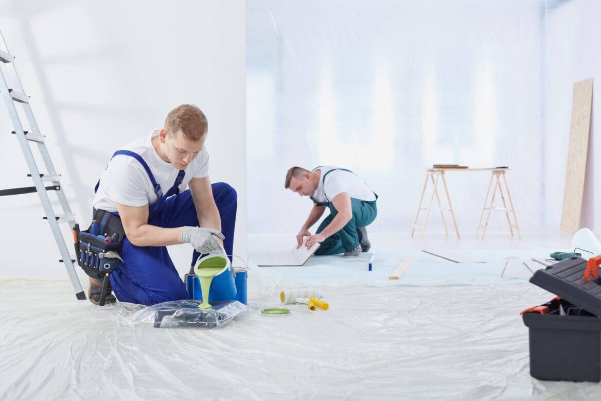 Two workers in a bright room; one pours green paint into a tray, the other measures wood. Tools and ladder nearby, evoking teamwork and focus.